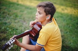 Photo of a child with hearing aid playing guitar while sitting on the grass